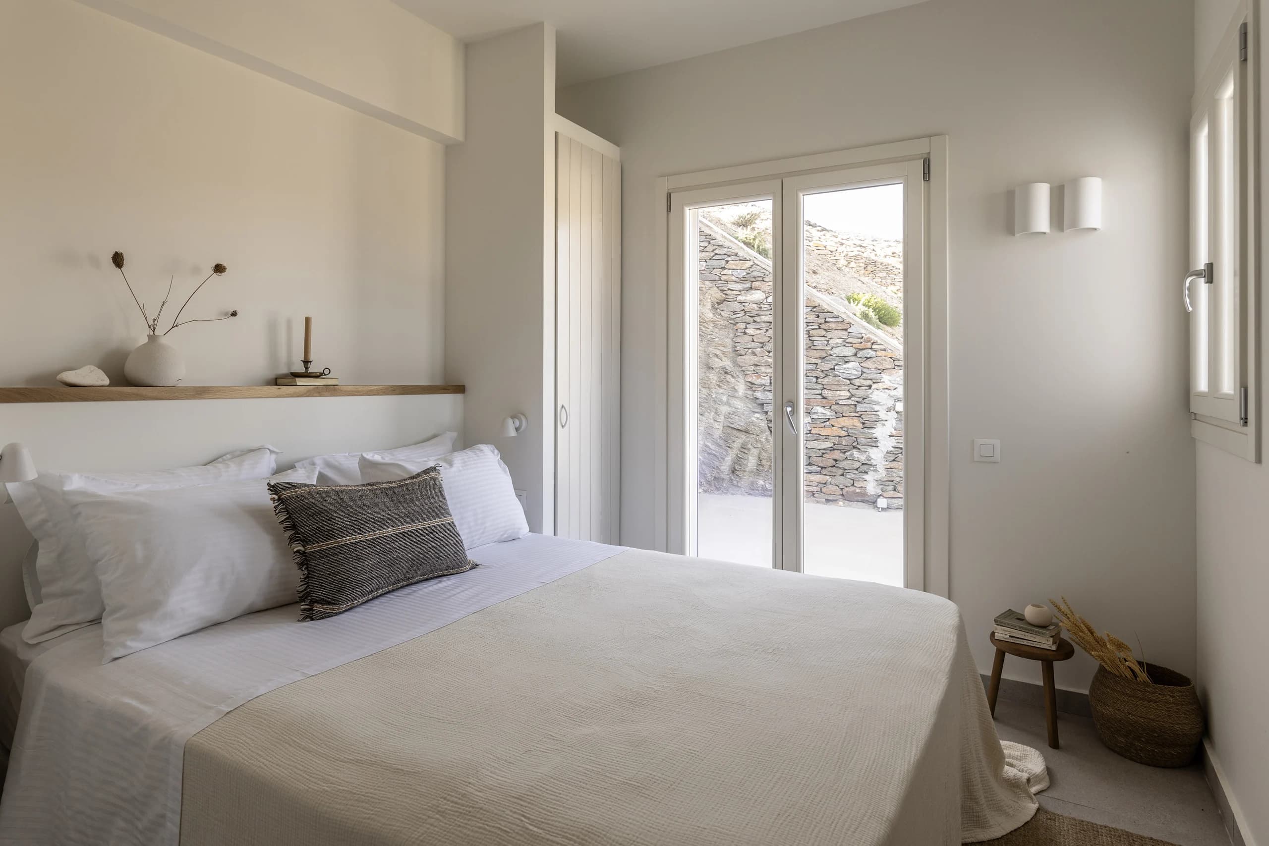 A serene bedroom with a neatly made double bed, soft beige bedding, and white pillows. A wooden shelf holds minimalist decor, and glass doors open to a stone-paved outdoor area with natural light pouring in.