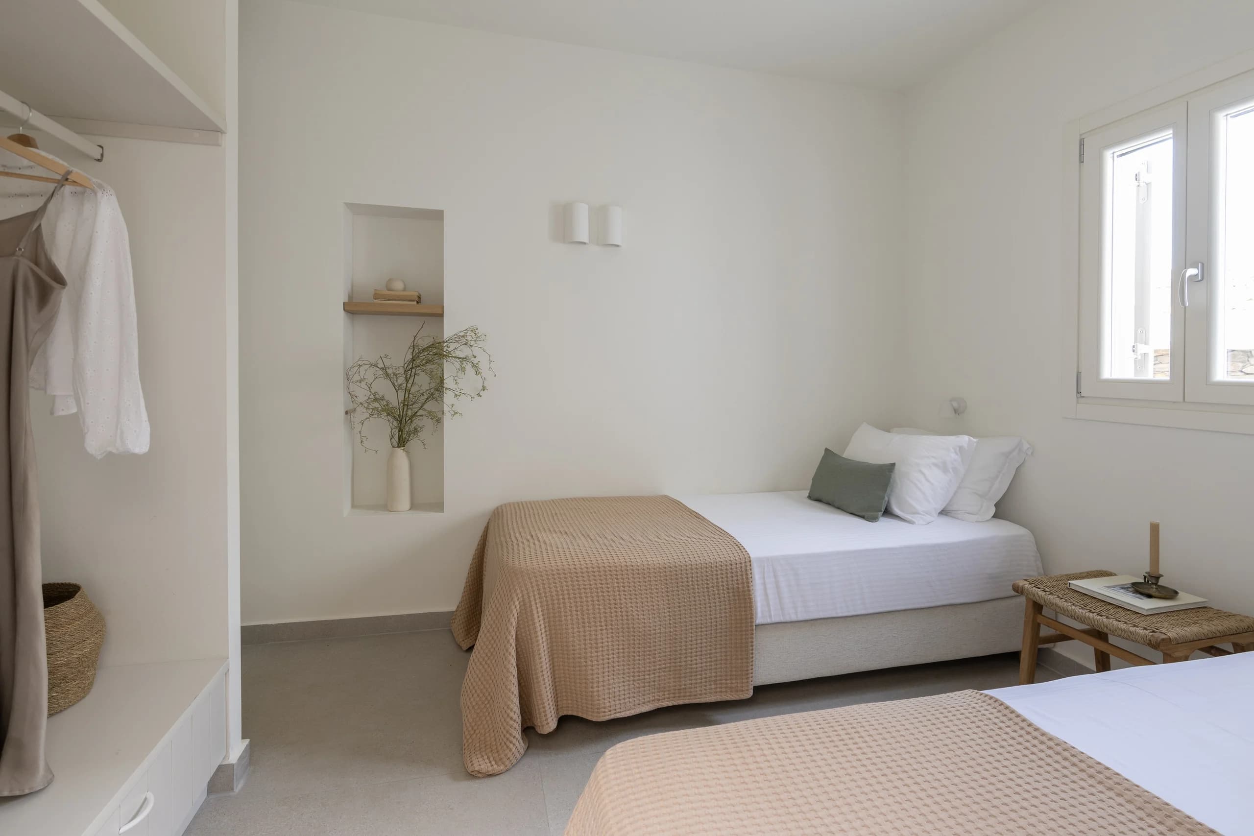 A bright bedroom with two single beds featuring beige bedspreads and white pillows. The room includes built-in shelves with minimalist decor and a simple, calming design.