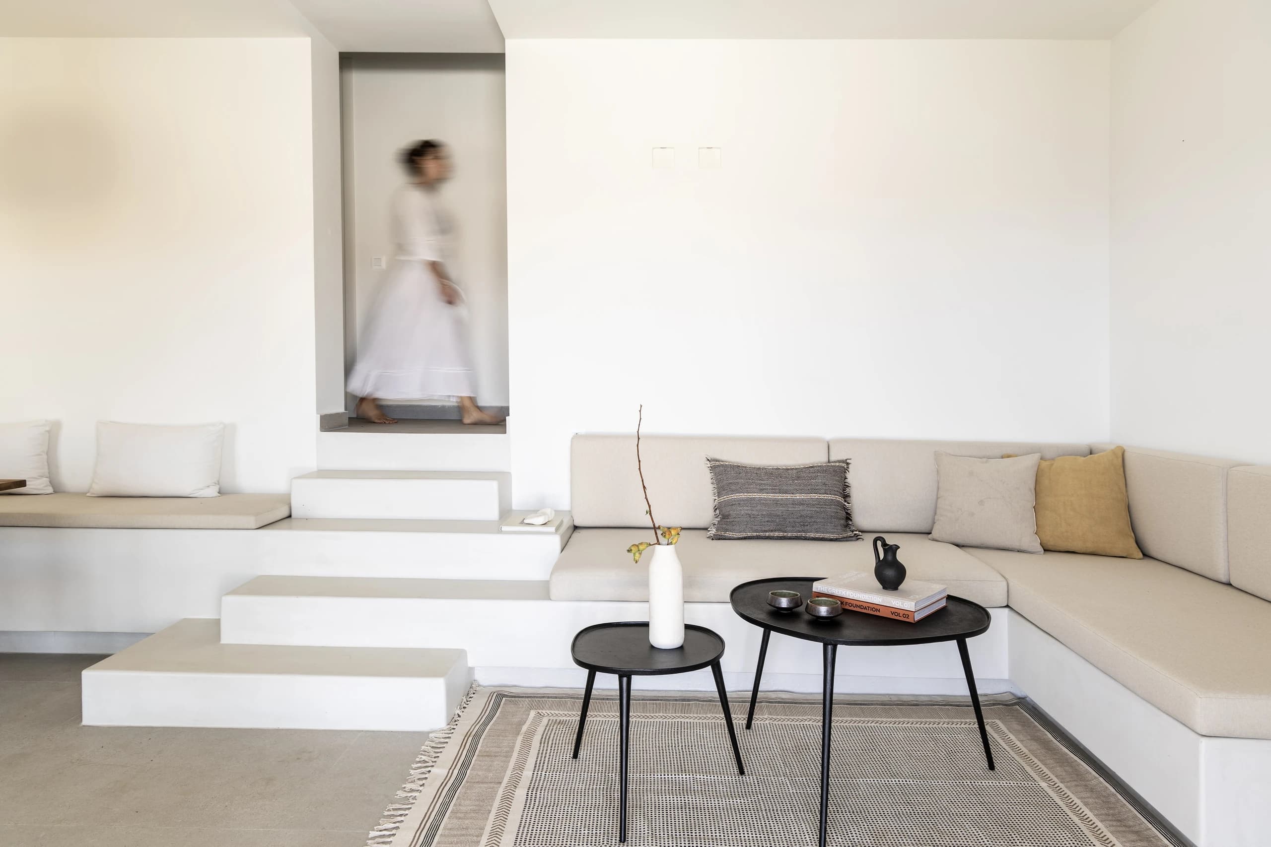 A built-in bench with neutral cushions and a modern black coffee table in the living area. A staircase leads to another part of the house, and a person in a white dress walks by, creating a dynamic and inviting ambiance.