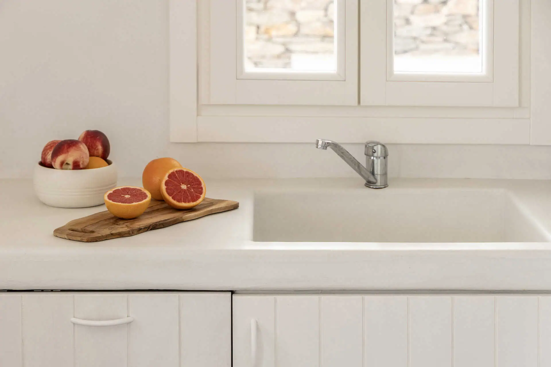 Close-up of a kitchen counter with fresh grapefruits on a wooden cutting board and a bowl of apples, placed next to a white sink and window with natural light streaming in.
