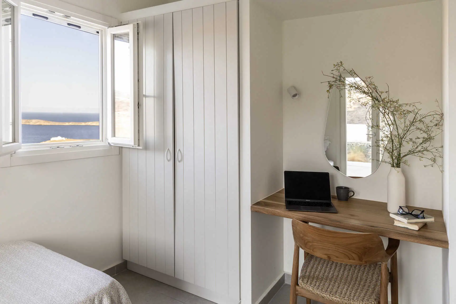 A bright and cozy bedroom corner with a wooden desk, a laptop, and decorative accents. An open window offers a stunning view of the Aegean Sea, while a built-in wardrobe provides ample storage space.