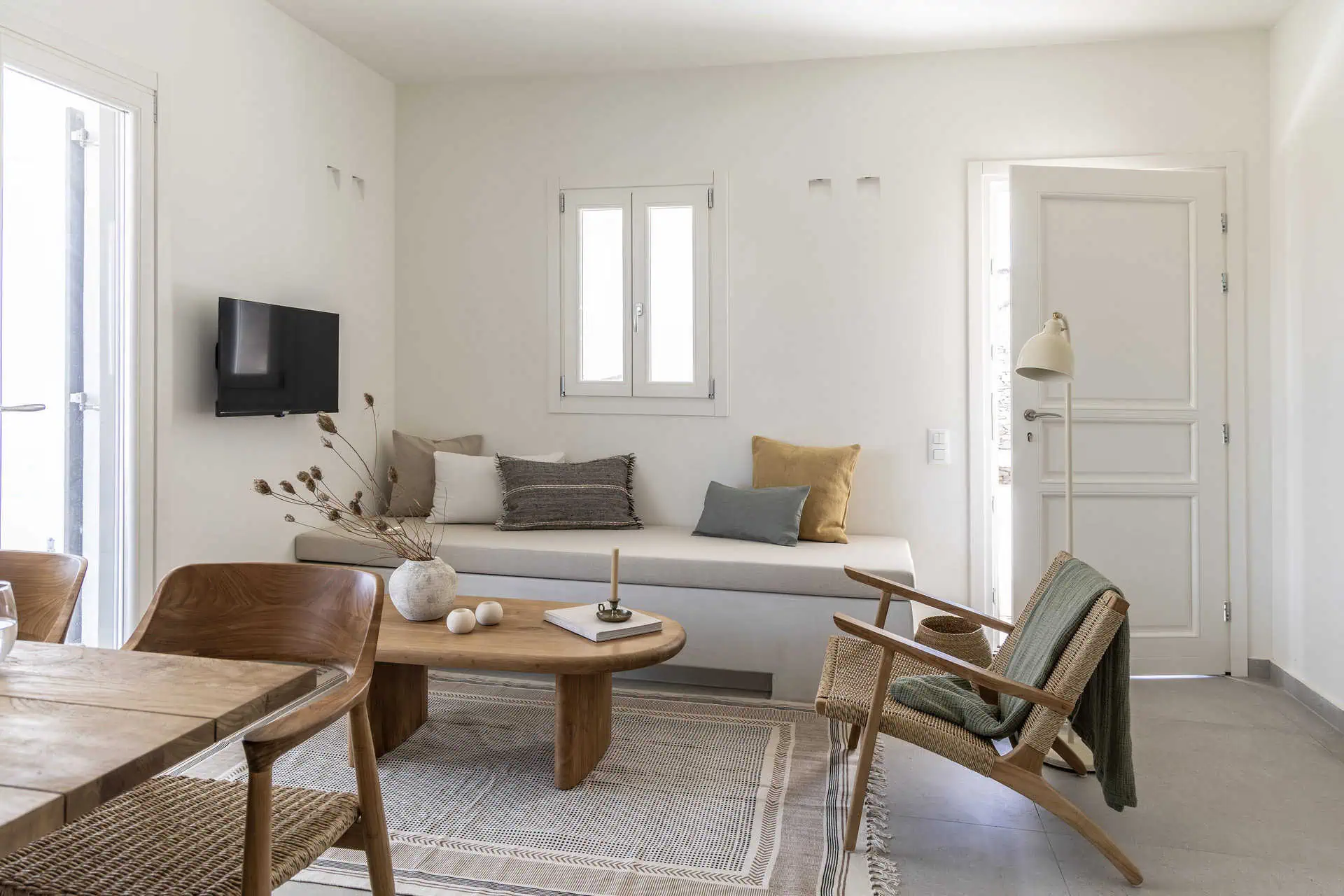 A cozy living room featuring a built-in sofa with decorative cushions, a wooden coffee table, and natural woven chairs. The minimalist decor is complemented by soft lighting and a bright, airy ambiance.