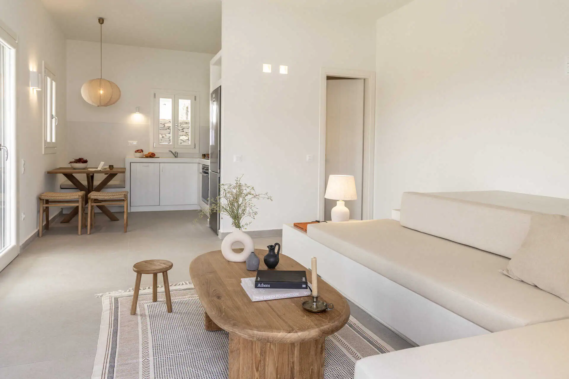 Bright and cozy living space with a minimalist beige sofa, wooden coffee table, and a view of the open kitchen and dining area, featuring natural light and soft decor elements.
