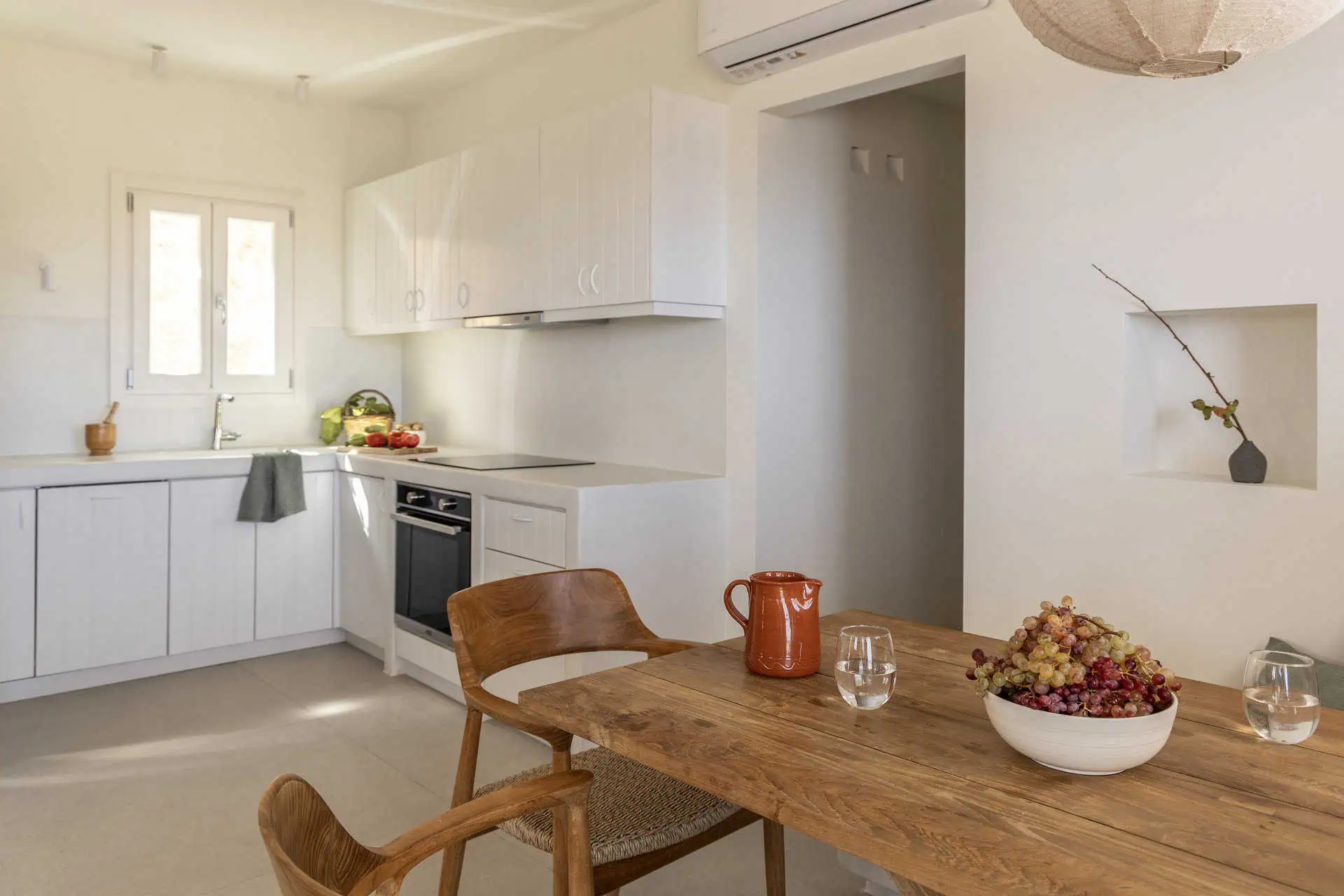 A bright and airy dining area with a wooden table and chairs, adjacent to a modern, fully equipped kitchen. Fresh fruit and vegetables add a warm, inviting touch to the minimalist decor.