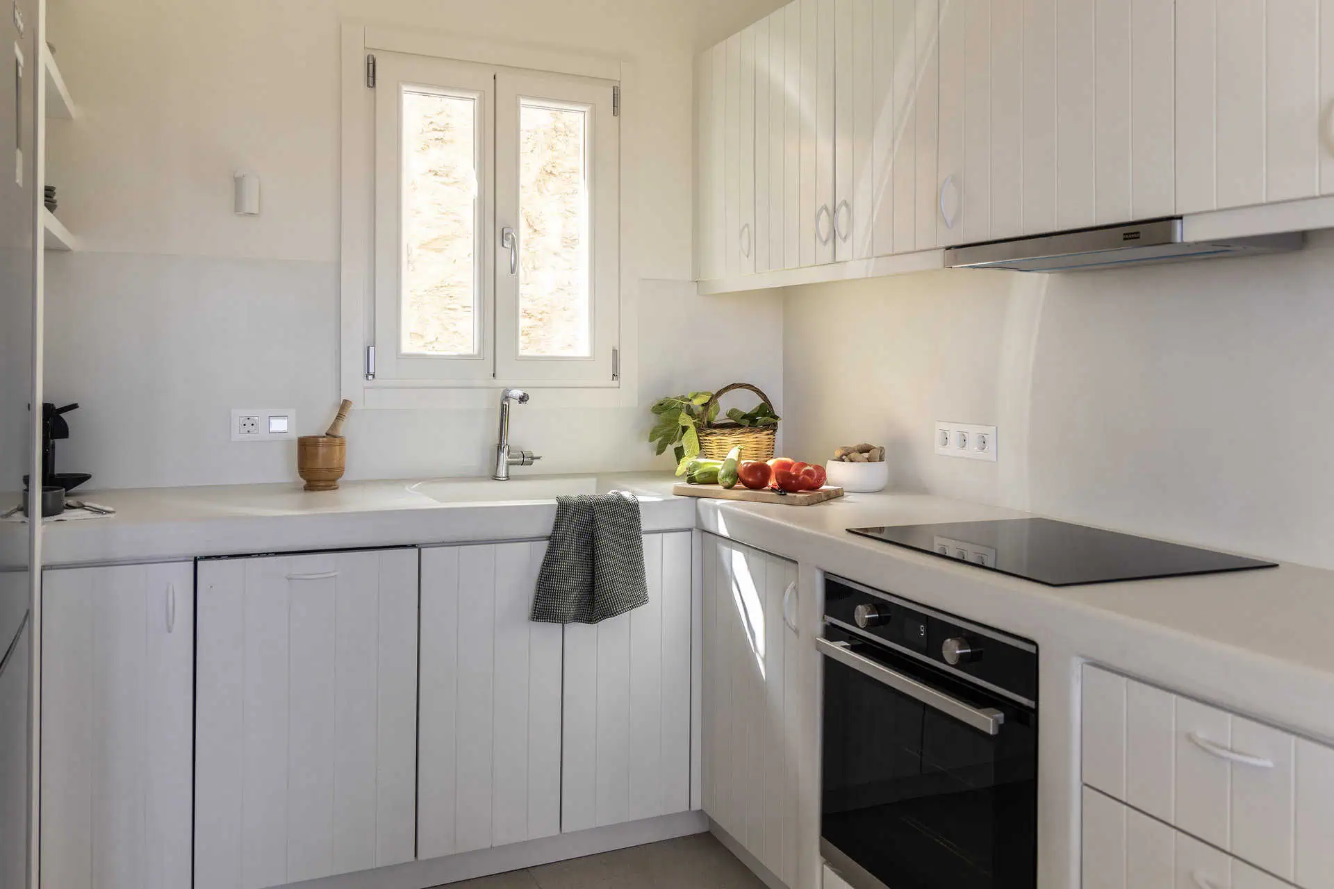A modern and fully equipped kitchen with sleek white cabinetry, a built-in oven, and a bright window letting in natural light. Fresh vegetables and herbs are arranged on the counter, adding a touch of warmth and vibrancy.
