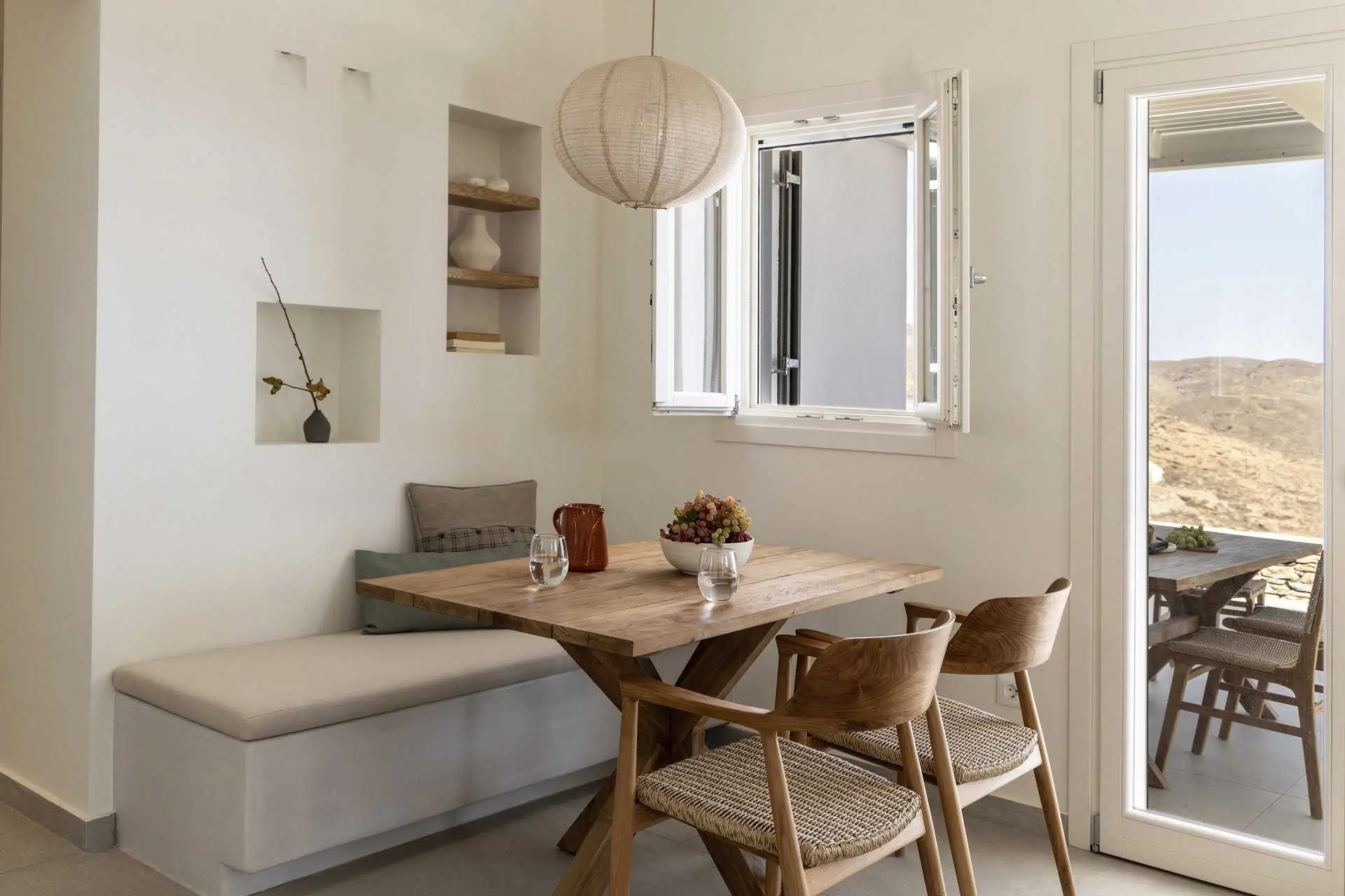 A cozy indoor dining nook featuring a wooden table with chairs and a built-in bench. Soft cushions, a pendant light, and open shelving with minimalist decor add charm, while an open window offers natural light and views of the surrounding landscape.