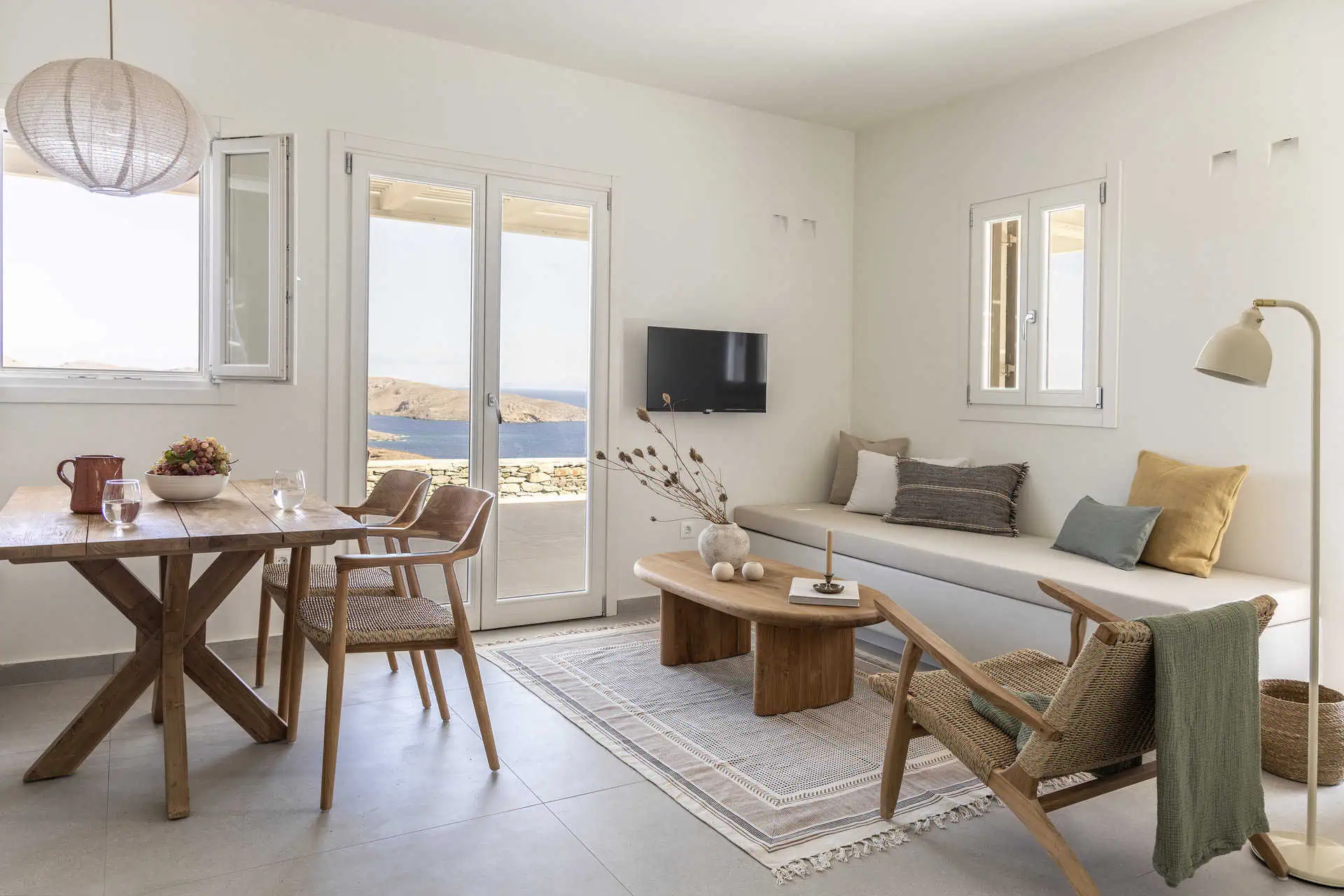 A bright and inviting living and dining space with wooden furniture, cozy cushions, and natural decor. The room features large windows and doors that open to stunning views of the Aegean Sea, creating a serene and airy atmosphere.
