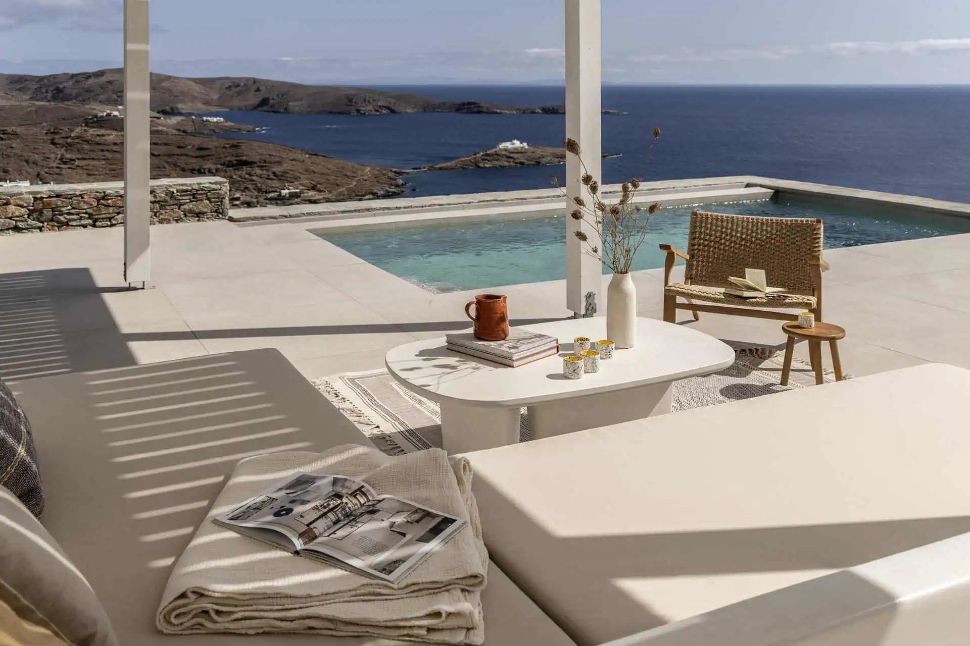 A luxurious outdoor lounging area with a private pool overlooking the Aegean Sea. The space features a comfortable daybed, a coffee table styled with books and decor, and a woven chair, creating a serene and elegant atmosphere.