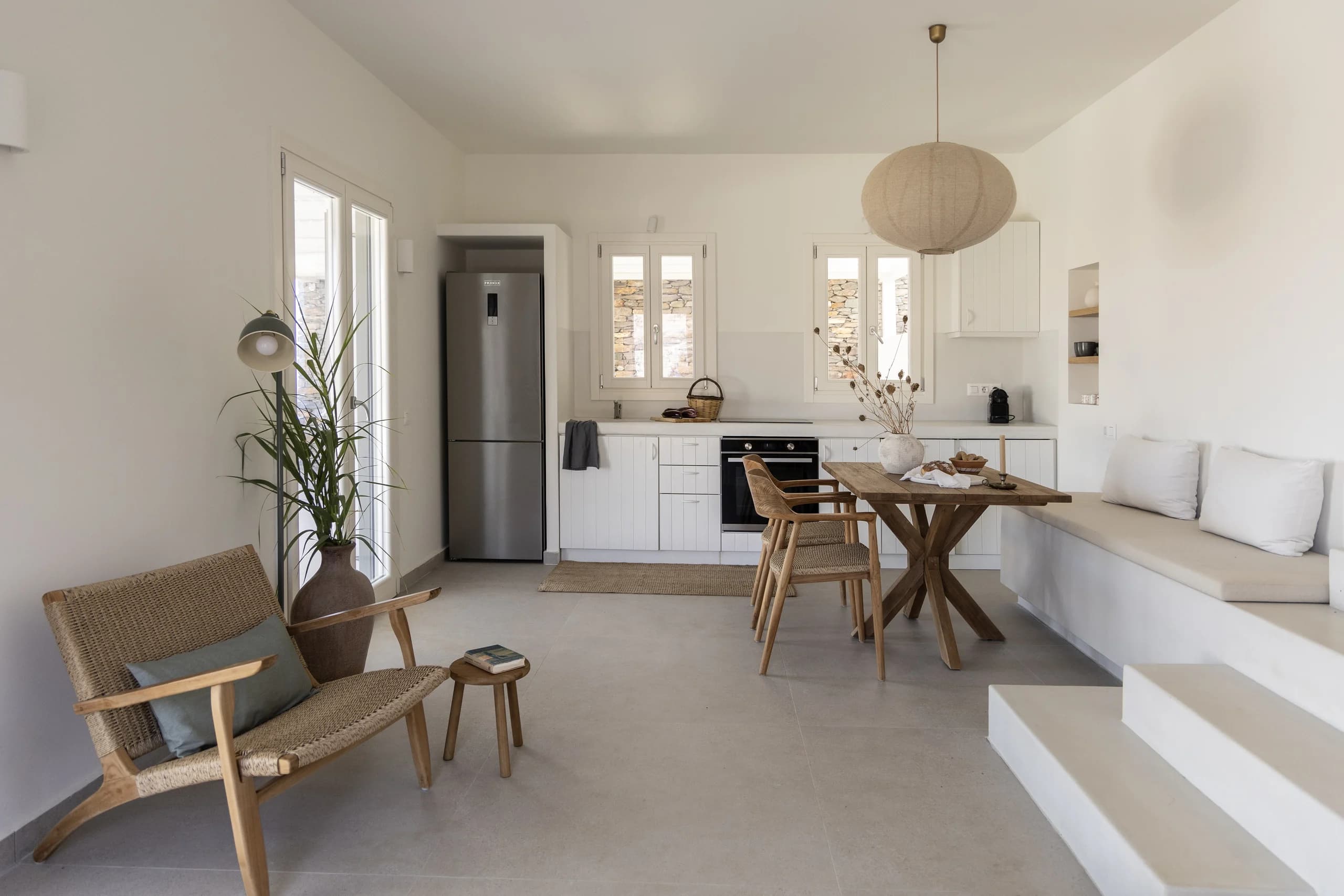 A close-up view of the open-plan living and kitchen area, showcasing the natural wooden furniture, woven chairs, and integrated bench seating with soft white cushions. A potted plant adds greenery to the serene space.