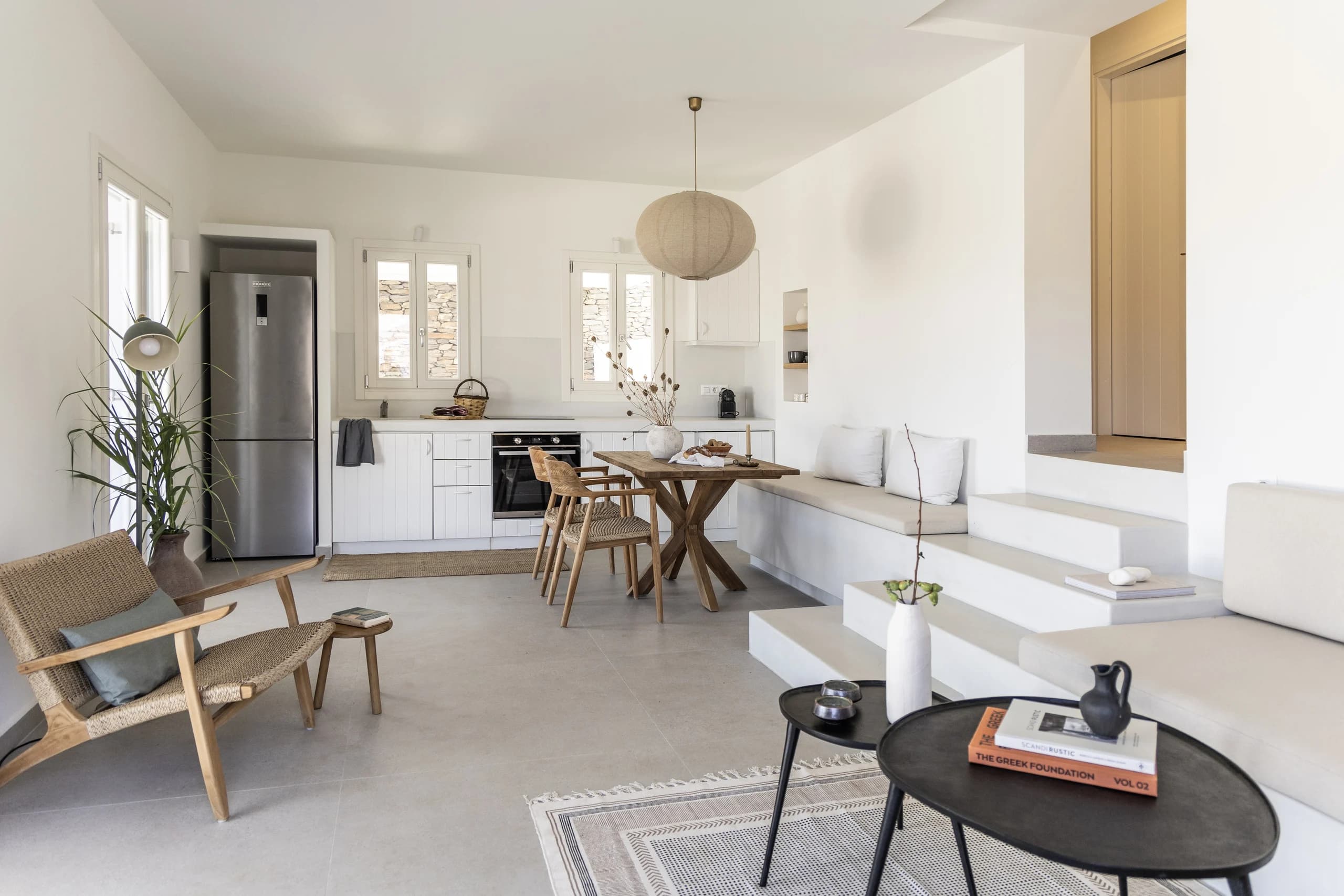 A cozy open-plan living and kitchen area with a minimalist design. Features include a wood dining table with wicker chairs, a built-in bench with white cushions, a modern kitchen with white cabinets, and natural light streaming through large windows.