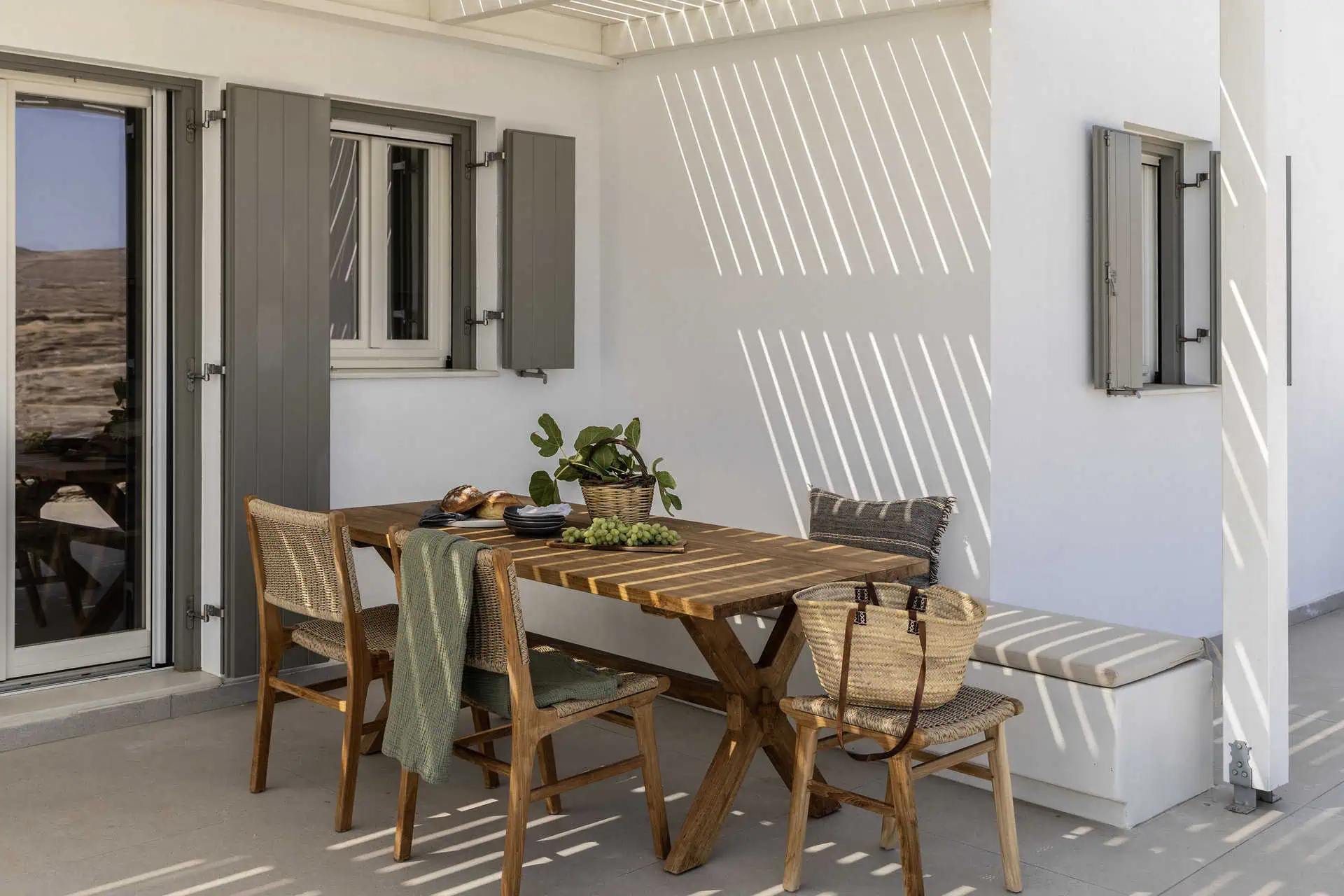 A charming outdoor dining area featuring a wooden table with woven chairs, set under a pergola casting patterned shadows. The space is adorned with a potted plant, fresh fruit, and cozy cushions, creating an inviting atmosphere for alfresco meals.