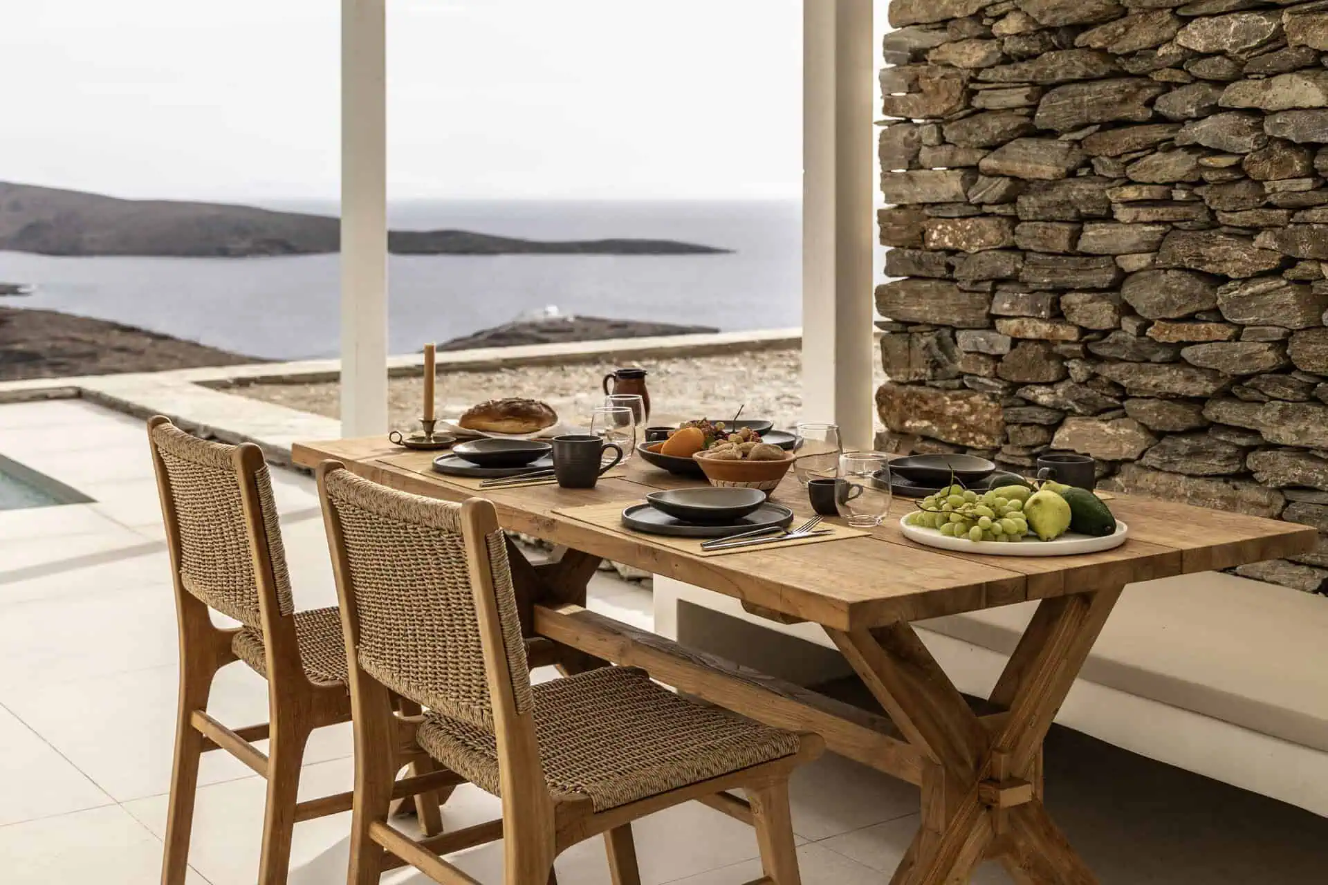 Outdoor dining setup with a wooden table featuring fresh fruits, bread, and tableware, overlooking the Aegean Sea and surrounded by rustic stone walls.