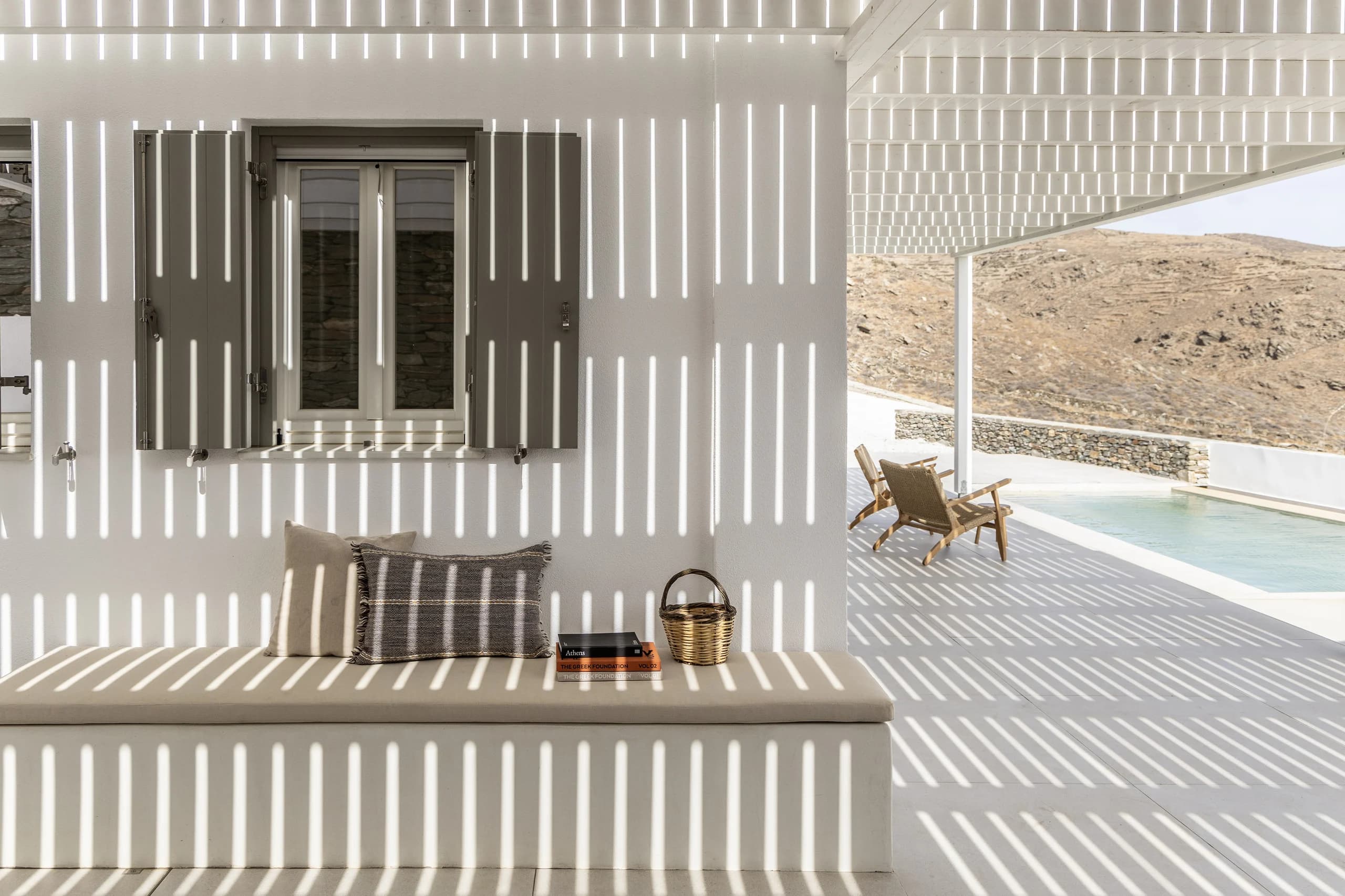 An outdoor seating area with a built-in bench and decorative pillows under a pergola casting striped shadows. A natural stone wall complements the view of the surrounding landscape.