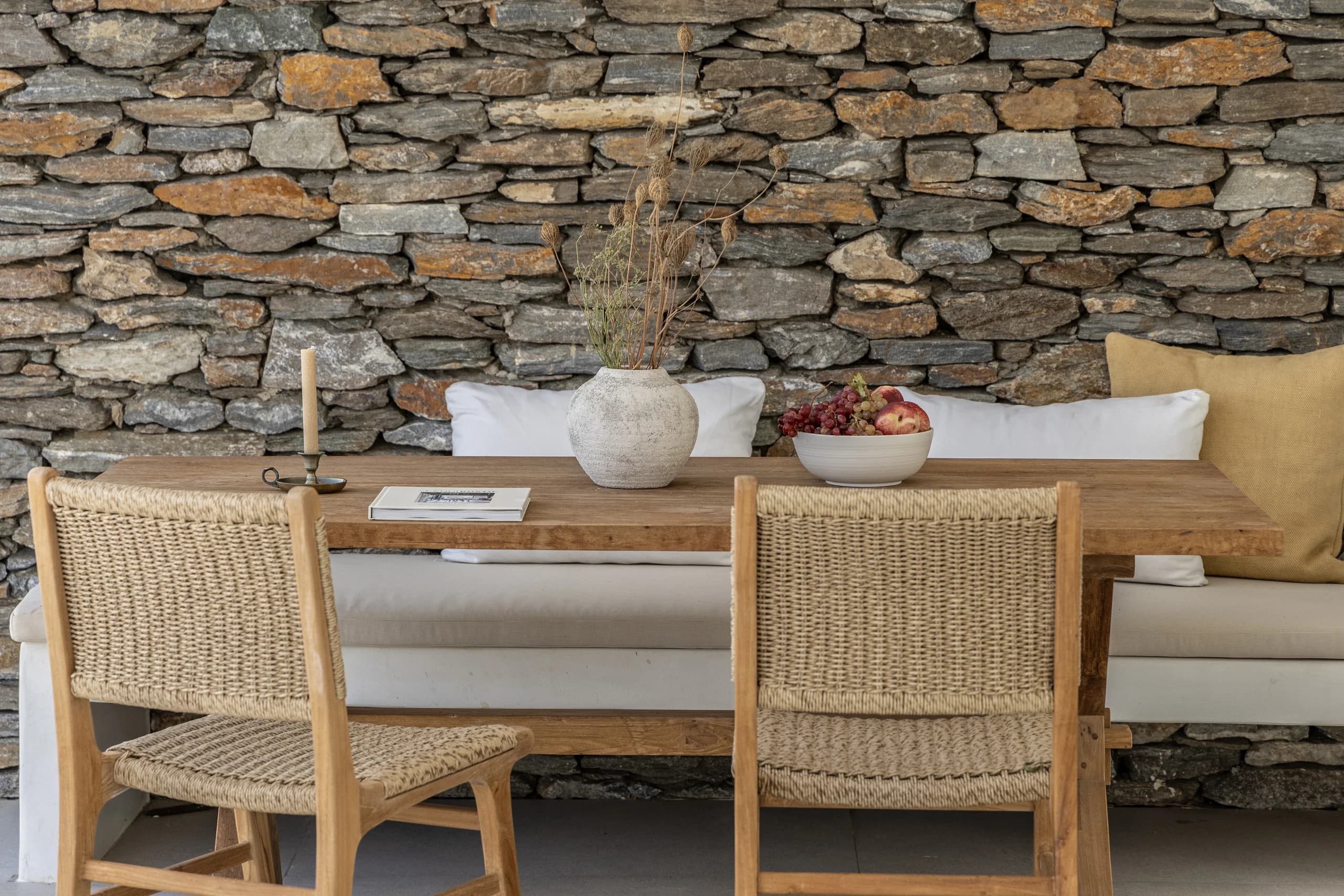 A close-up of the rustic outdoor dining table with a bowl of fruit, a candle, and a textured ceramic vase. The stone wall adds warmth and character to the cozy setting.