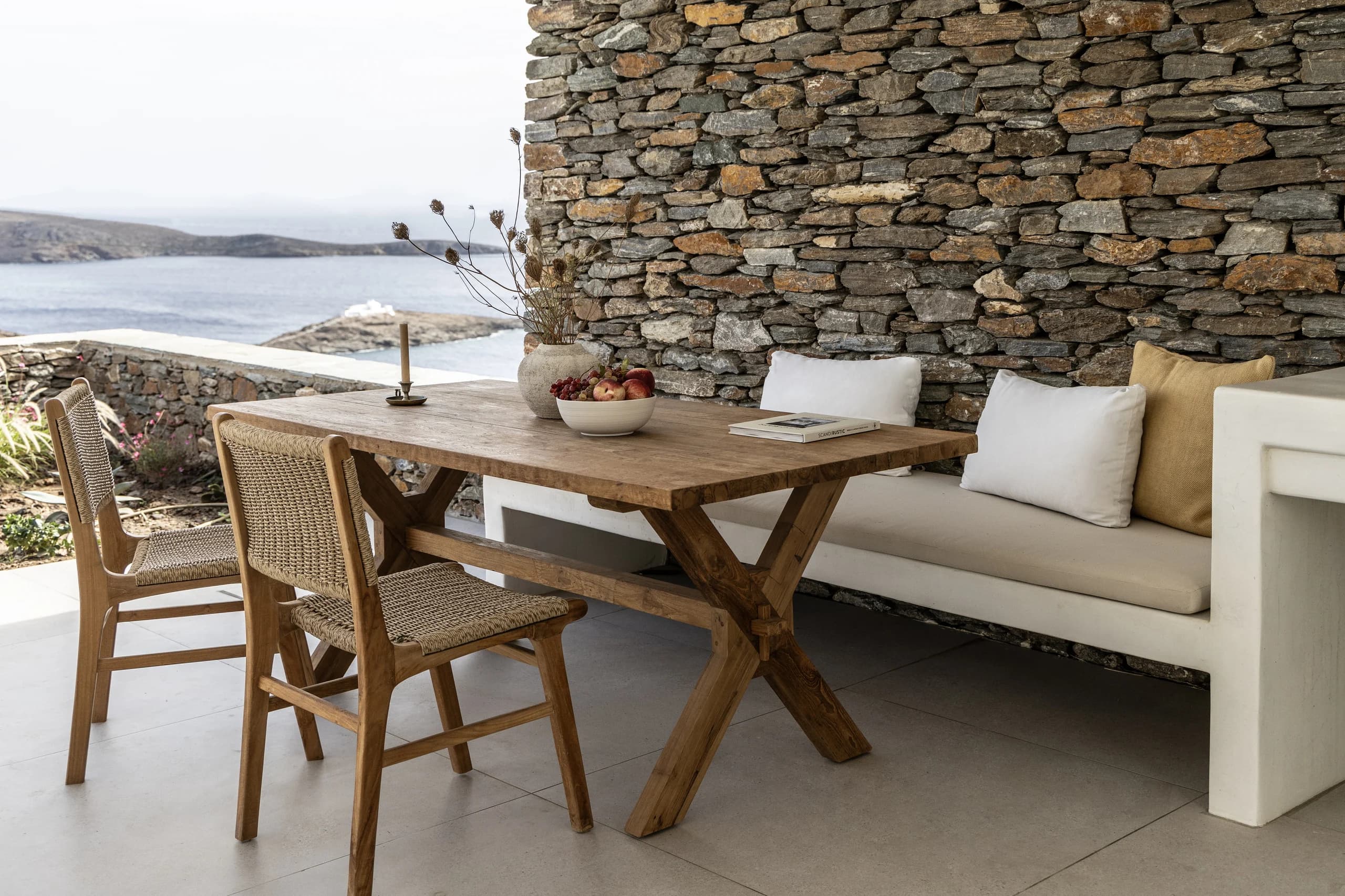 A rustic outdoor dining area featuring a wooden table, woven chairs, and a built-in bench with white cushions. The natural stone wall provides a backdrop, and the table is adorned with decorative vases and fruit.
