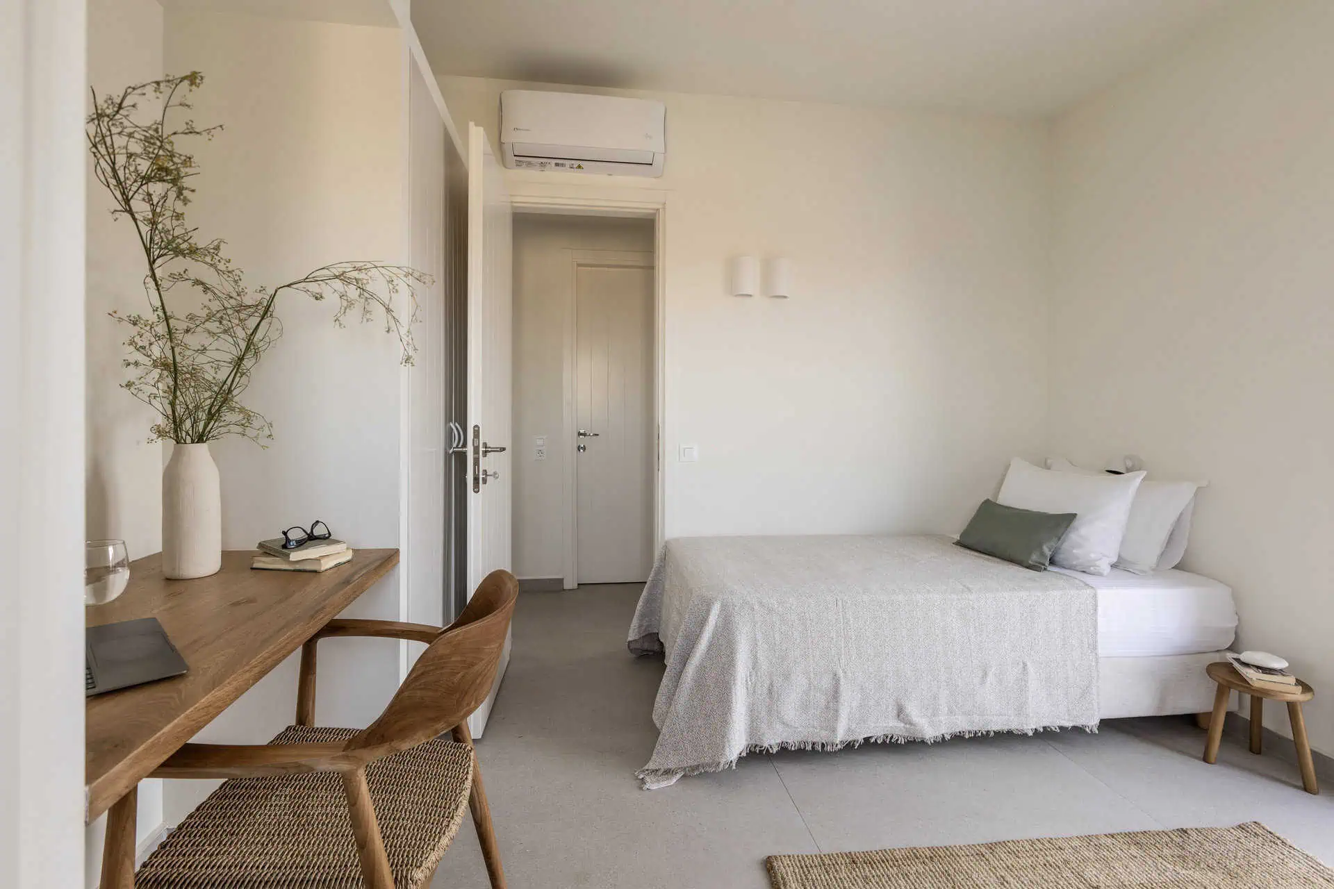 A serene bedroom with a neatly made bed, a cozy workspace featuring a wooden desk and chair, and minimalist decor. The room is bright and airy, with neutral tones and an air conditioner for added comfort.