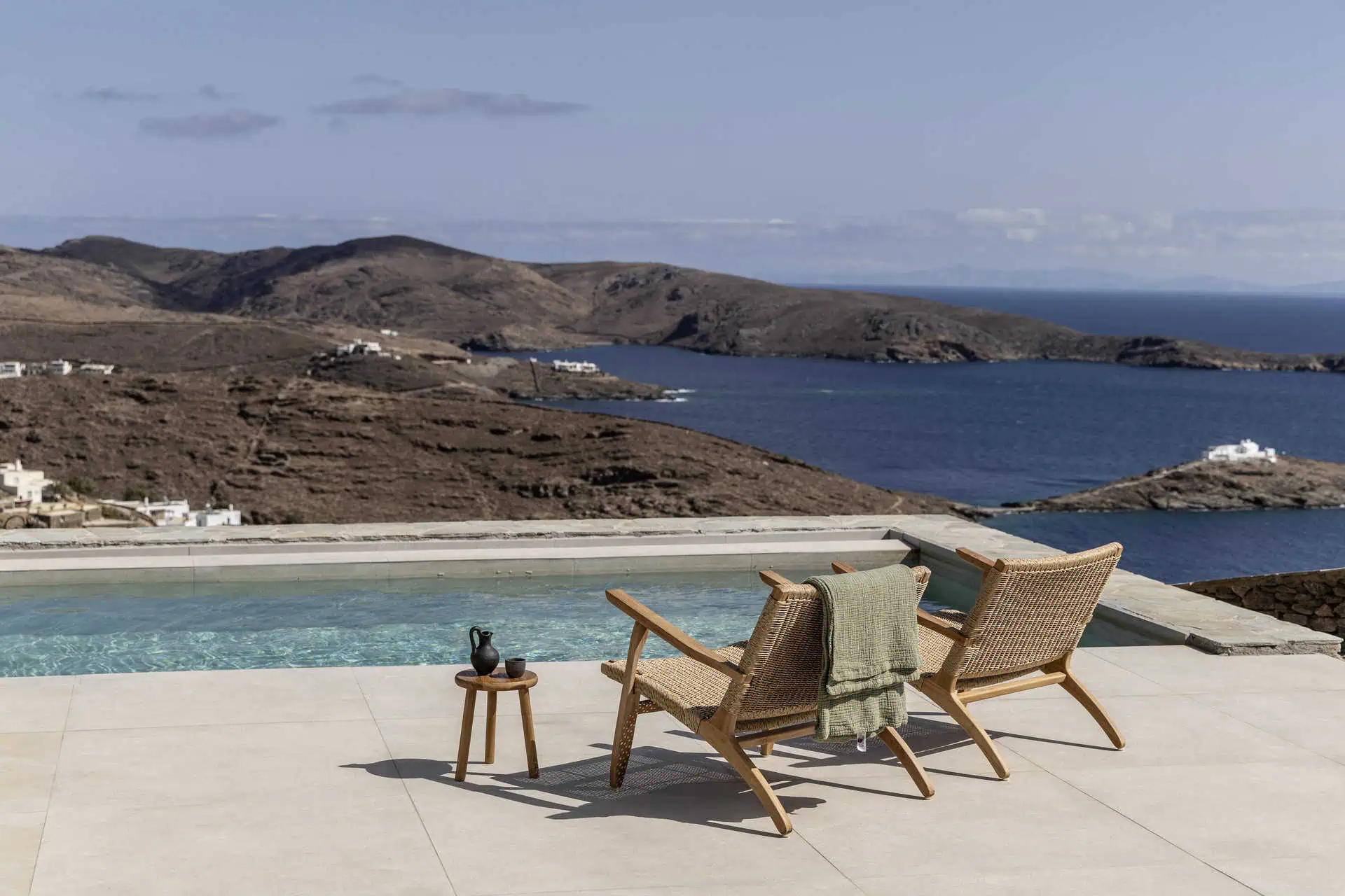 A serene outdoor pool area overlooking the Aegean Sea and rugged hills. Two wooden lounge chairs with woven backs and a small side table create the perfect spot for relaxation in this picturesque setting.