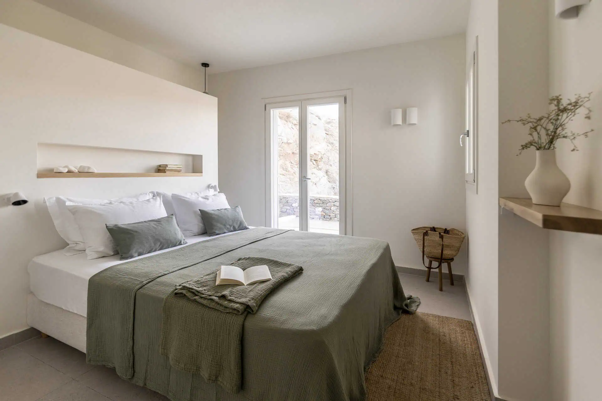 A bright and stylish bedroom with a large bed dressed in soft green linens and white pillows. The room features minimal decor, a built-in shelf, and double doors leading to an outdoor space, creating a cozy yet airy atmosphere.