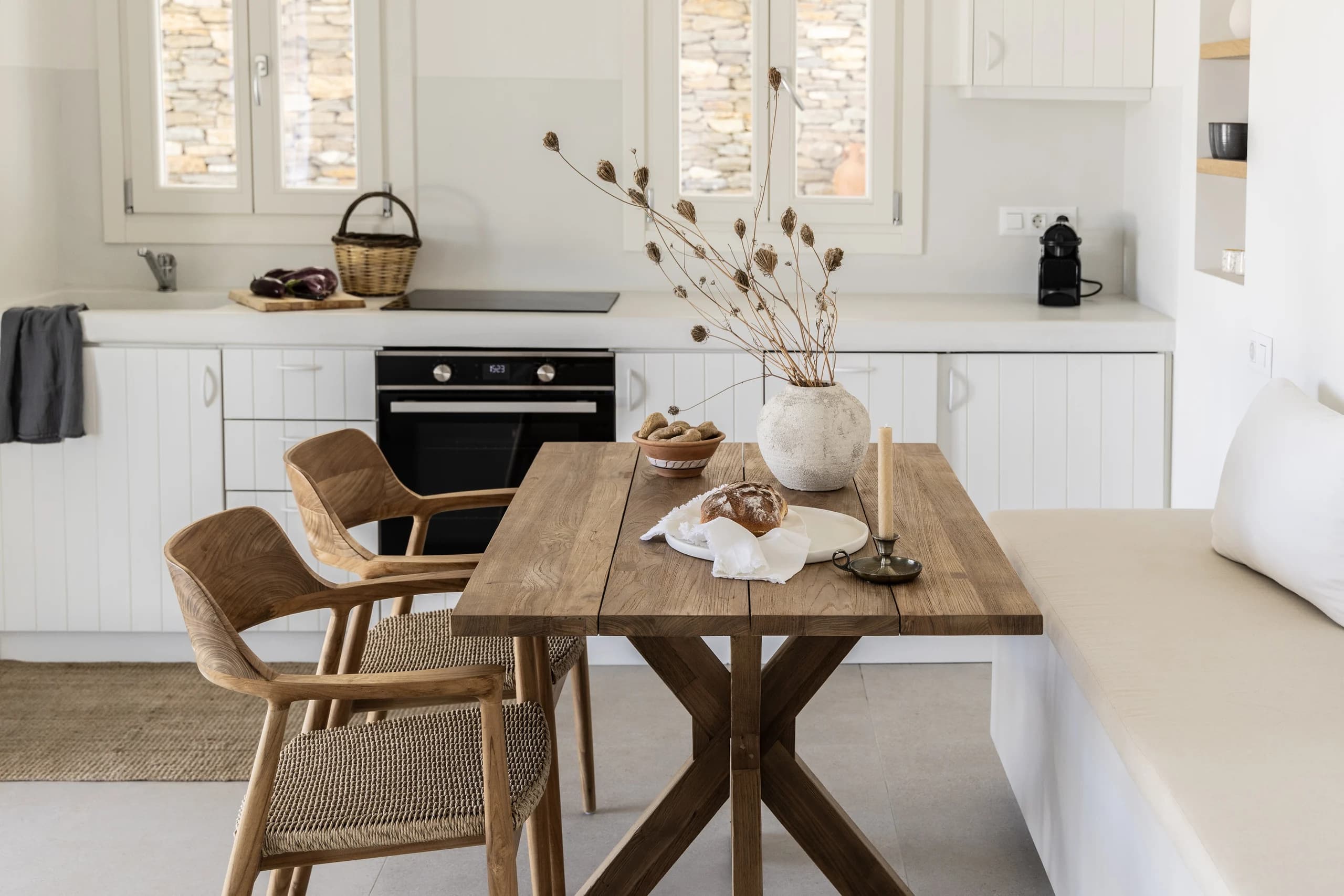 A close-up of the wooden dining table set for a meal with decorative vases, dried branches, bread, and fruit. The white kitchen in the background is minimalistic and functional, with natural stone accents visible through the window.
