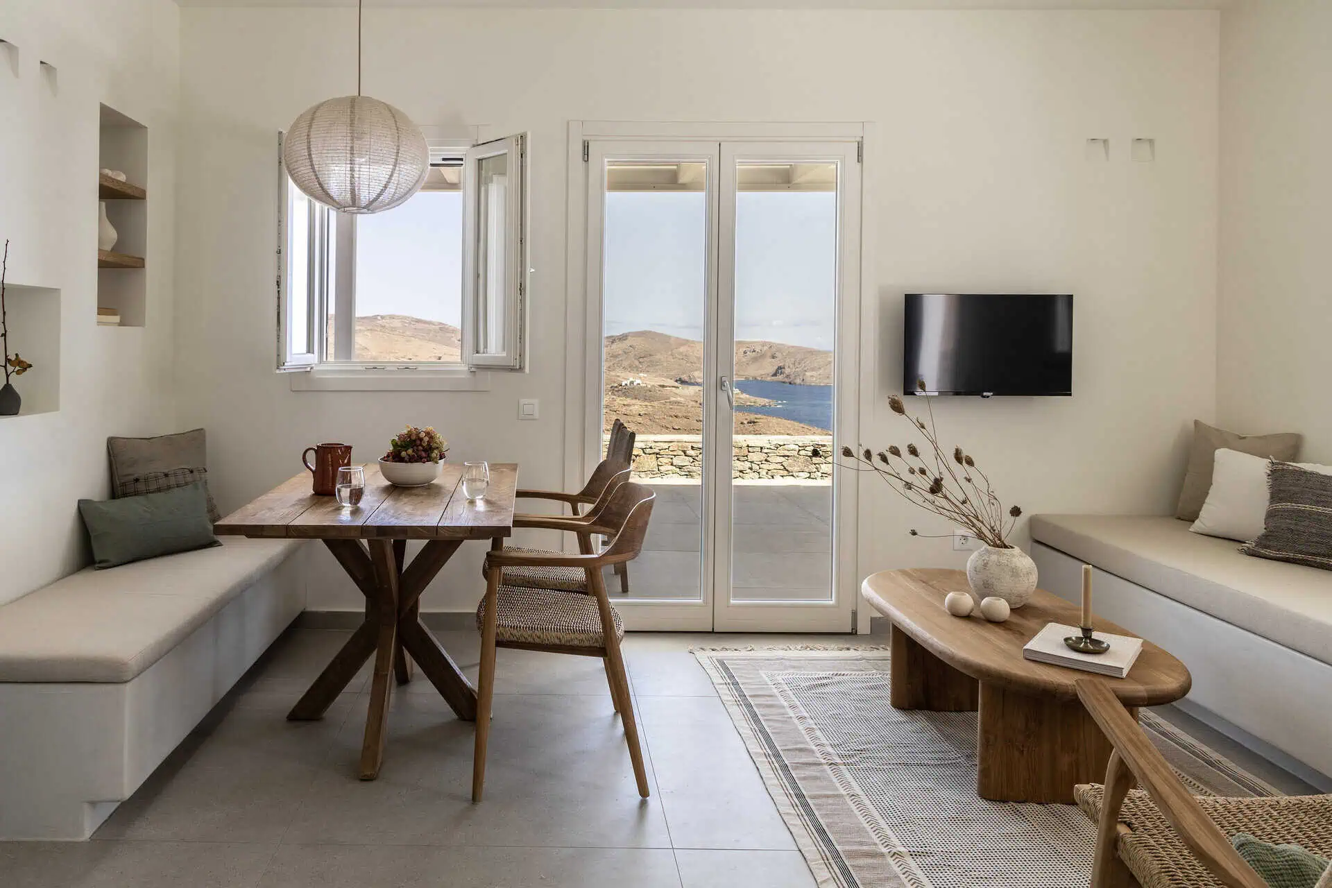 A cozy living and dining area with a wooden table, built-in bench seating, and a comfortable sofa. The space is decorated with natural tones, minimalist accents, and features large glass doors offering stunning views of the surrounding landscape and sea.