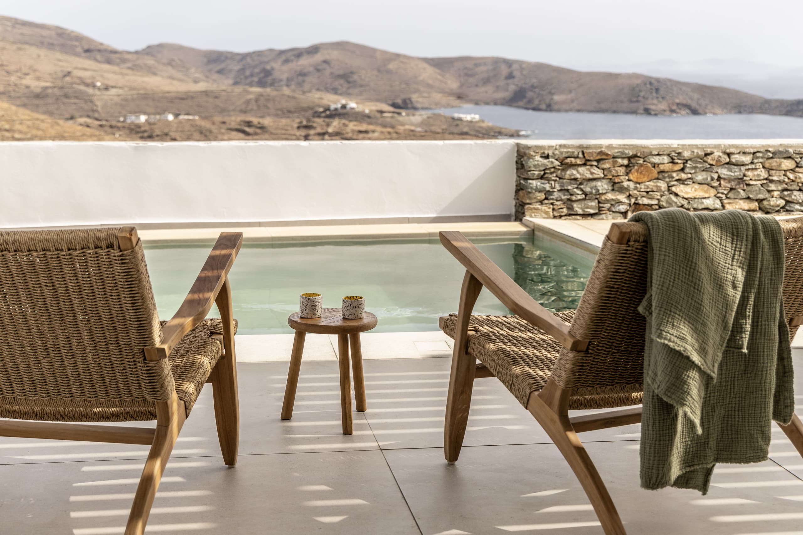 Two woven wooden chairs facing a serene private pool and the rolling hills of Kythnos, with a green towel draped over one chair and ceramic mugs on a small wooden table.