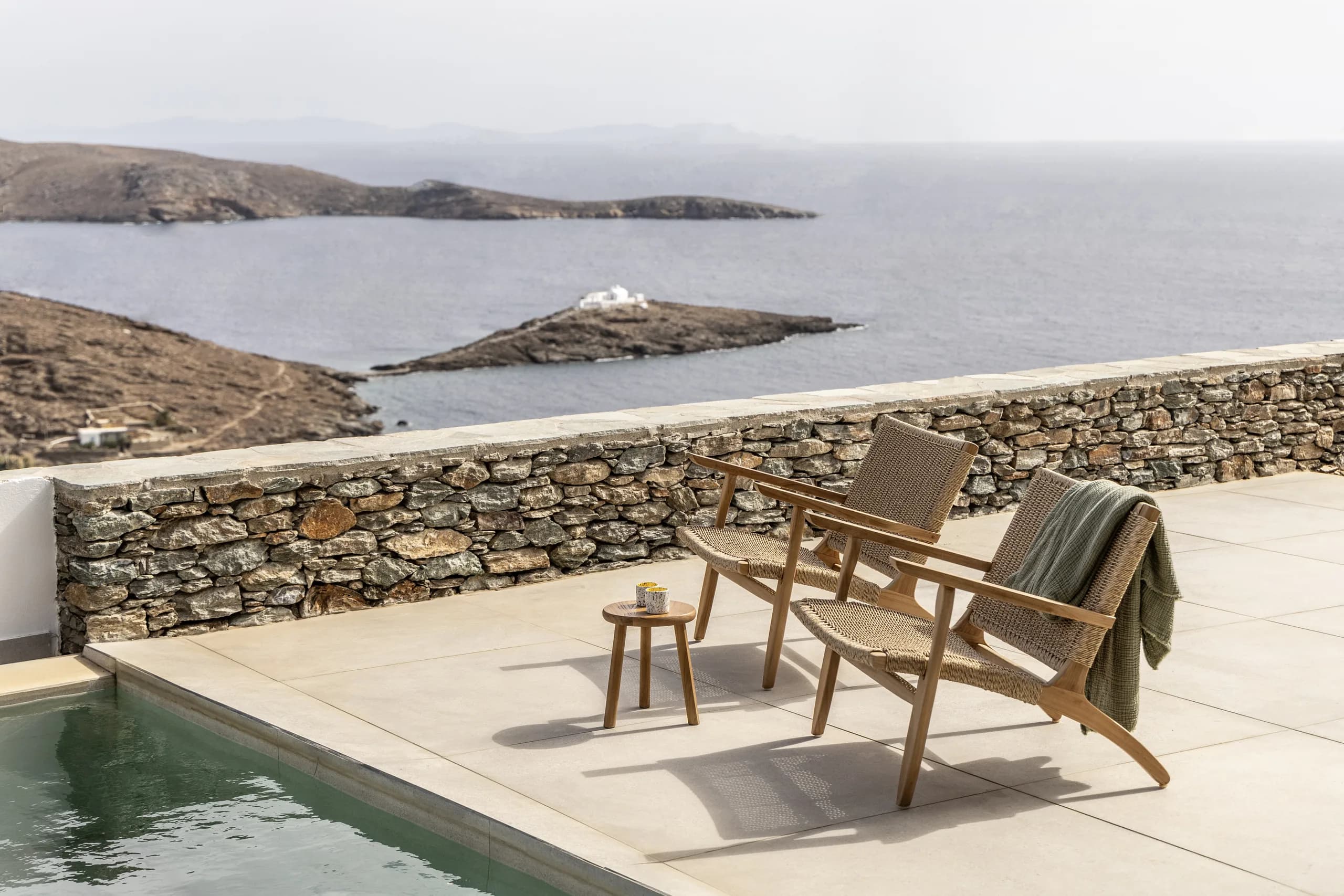 An outdoor space with woven chairs and a small wooden table by a pool, overlooking a stunning view of the sea and nearby islands. The scene is peaceful and inviting.